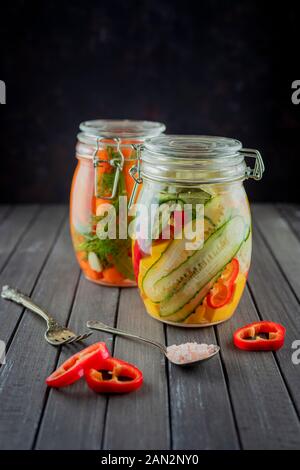 due vasi di vetro di cetrioli fermentati, carote, pepe, sale, verdure su fondo scuro. fermentazione è una fonte di probiotici. vegetari popolari Foto Stock