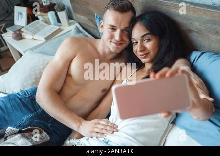 Razza mista giovane. Giovane uomo e donna a casa sdraiato sul letto tenendo felice selfie sullo smartphone Foto Stock