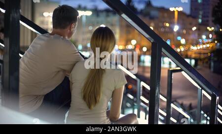 Giovane seduto su scale, avvolgente, guardando la sera illuminata città, amore Foto Stock