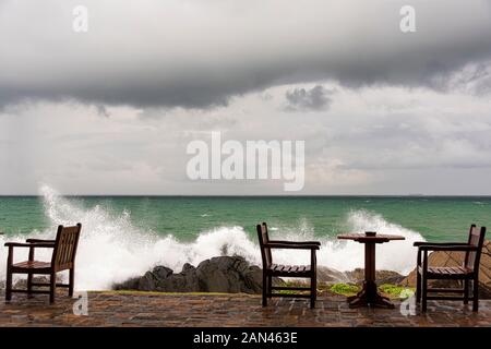 Galle, Sri Lanka, Settembre 2015: arrabbiato onde infrangersi sulle rocce della costa vicino abbandonato i tavoli e le sedie durante una tempesta estiva Foto Stock