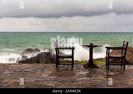 Galle, Sri Lanka, Settembre 2015: arrabbiato onde infrangersi sulle rocce della costa vicino abbandonato i tavoli e le sedie durante una tempesta estiva Foto Stock