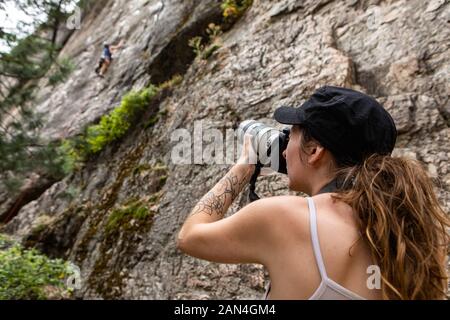 Un basso angolo di vista come una donna usa una telecamera professionale con lenti molto grandi di fotografare un uomo salire una ripida rupe in natura, visto sfocata in background Foto Stock