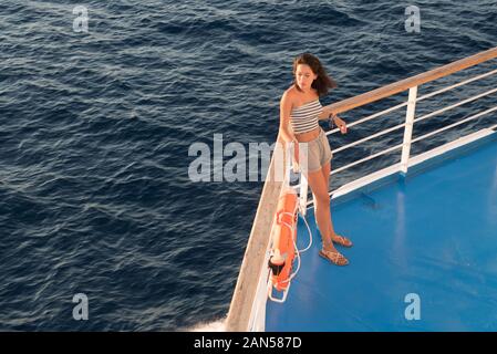 Asian teen nel tubo superiore e shorts in piedi sul ponte blu accanto a orange salvagente sul traghetto andando a Lesbo Foto Stock