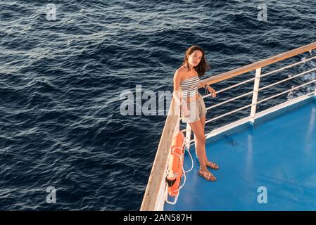 Asian teen nel tubo superiore e shorts in piedi sul ponte blu accanto a orange salvagente sul traghetto andando a Lesbo Foto Stock