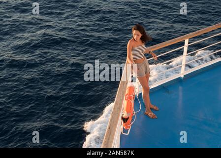 Asian teen nel tubo superiore e shorts in piedi sul ponte blu accanto a orange salvagente sul traghetto andando a Lesbo Foto Stock