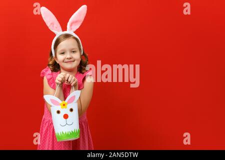 Bella carino bambina in Easter Bunny orecchie contiene un sacchetto per i dolci e le uova. Ritratto di un bambino su uno sfondo di colore rosso. Copia dello spazio. Foto Stock