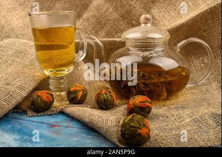 Fiore di infuso di tè in una teiera in vetro, un bicchiere di tè di fiori e palline di tè di fiori su uno sfondo di homespun ruvida stoffa. Close up Foto Stock