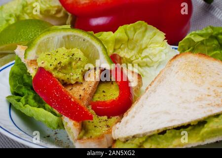 Piastra con sandwich vegan, pasta di avocado. Pezzi di peperone rosso, lattuga romana e limone. Colazione dietetica. Foto Stock