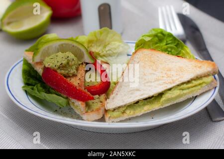 Piastra con sandwich vegan, pasta di avocado. Pezzi di peperone rosso, lattuga romana e limone. Colazione dietetica. Foto Stock