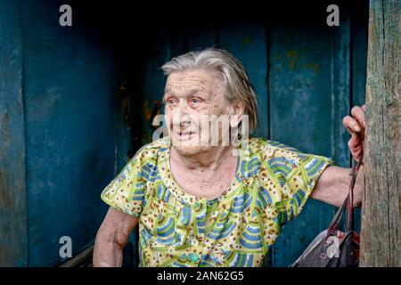 Chelyabinsk, Russia - Maggio 2019: Una donna anziana si appoggia su una tavola di legno stipite della porta in una vecchia casa. Foto Stock