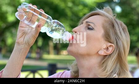 Donna matura acqua potabile dalla bottiglia nel parco, mantenendo il bilancio idrico Foto Stock