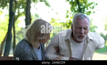 Senior sofferenza uomo forte dolore alla schiena, passeggiate nel parco, caring moglie aiutandolo Foto Stock