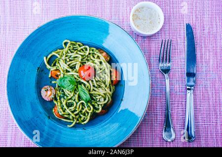 In casa italiana pici pasta verde con pesto e pomodori ciliegia sulla piastra blu con parmigiano e posateria Foto Stock