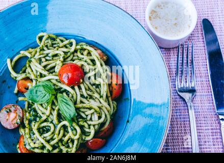 In casa italiana pici pasta verde con pesto e pomodori ciliegia sulla piastra blu con parmigiano e posateria Foto Stock