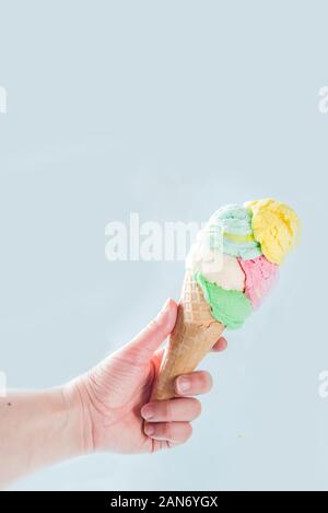 Pila di pastello colorato ice cream scoop nel cono di cialda, isolata su fondo azzurro. Donna mano trattiene grande cono gelato con sapore diverso Foto Stock