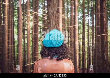 Donna che indossa una giacca di pelle marrone e verde piana-round hat guardando la foresta di alberi Foto Stock