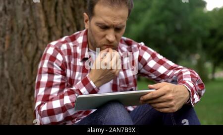 Concentrati gli uomini adulti utilizzando tablet nel parco, leggere l'articolo sulla salute mens Foto Stock