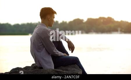 Malinconica adolescente di sesso maschile seduto da solo sulla pietra vicino al fiume e a pensare la vita Foto Stock