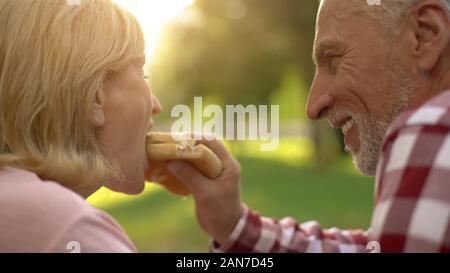 Uomo anziano alimentazione di sua moglie fast food Burger il picnic, data romantico in posizione di parcheggio Foto Stock