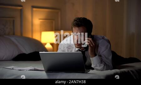 Uomo in tuta giacente sul letto lavorando su laptop e parlando al telefono, lifestyle occupato Foto Stock