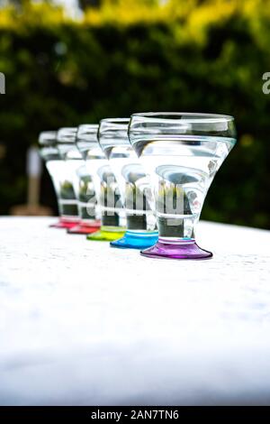 Colorato bicchieri su sfondo bianco in piedi in una fila nella luce naturale con fogliame verde. sfondo rosso, arancio, verde blu viola . Foto Stock