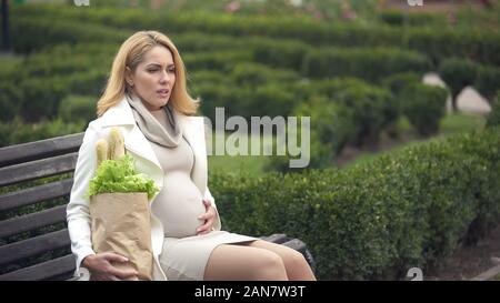 Stanco femmine gravide di seduta una panchina nel parco azienda sacchetto shopper, aspettandosi la fatica Foto Stock