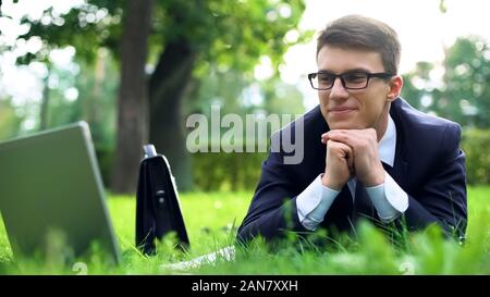Imprenditore giacente su erba, guardare video sul computer portatile e allegramente sorridente Foto Stock