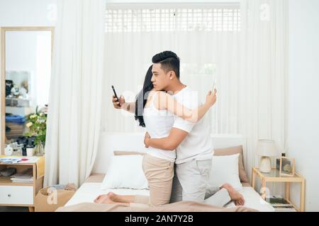 Coppia giovane in camera da letto. Vista laterale di infedele l uomo e la donna sono barare sul telefono mentre abbraccia ogni altro Foto Stock