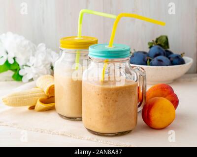 Banana pesche nettarine smoothie in due vasi, frutta fresca frappè su bianco tavola in legno rustico, vista laterale Foto Stock