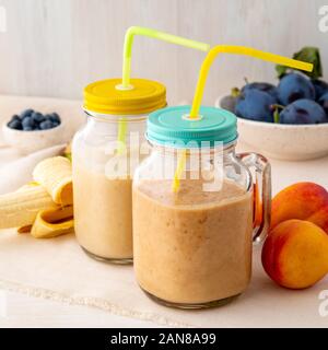 Banana pesche nettarine smoothie in due vasi, frutta fresca frappè su bianco tavola in legno rustico, vista laterale Foto Stock