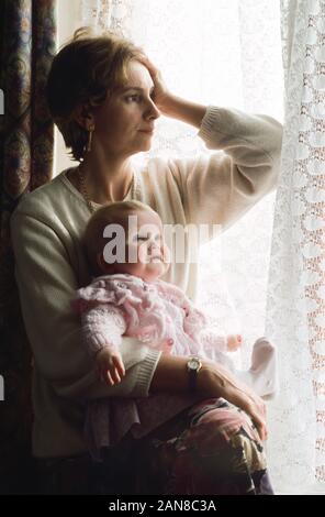 Giovane madre che soffrono di post natal depressione con il suo bambino seduto accanto a le porte del patio, guardando al di fuori sensazione triste Foto Stock