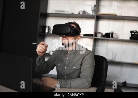 Giovane uomo barbuto seduto contro computer con occhiali per realtà virtuale cercando di toccare qualcosa o toccare qualche cosa di fronte a ciò che egli ha visto. Moderno Foto Stock