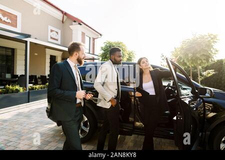 Salone auto, vendita e affitto concetto. Africani e uomini caucasici in business usura acquistare la vettura. Donna elegante vettura salesmanager mostra la vettura, apertura Foto Stock