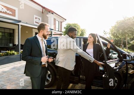 Clienti felici acquistare nuove auto in piedi nel salone auto all'aperto. Due uomini in abiti aziendali scegliendo una nuova vettura, mentre la giovane donna graziosa salesmanager Foto Stock