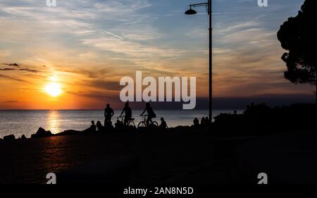 Drammatico e splendido tramonto a lungo la linea costiera del Mar Mediterraneo in Trieste Italia con sagome di un giovane e la gente seduta sulla spiaggia Foto Stock