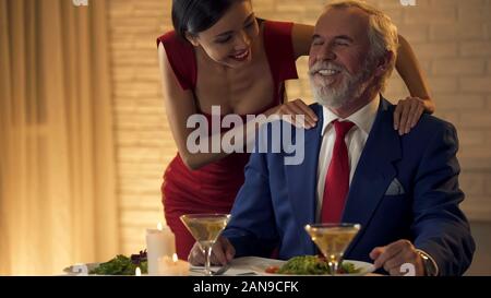 Giovane e bella signora massaggiare le spalle del vecchio uomo ricco, flirtare in data Foto Stock