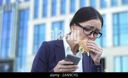 Giovane donna manager mangiare hamburger di malsano in fretta, controllo news sul telefono Foto Stock