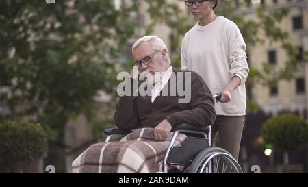 Giovane donna sulla passeggiata con triste pensionato in carrozzella, il caregiver assistenza, aiuto Foto Stock