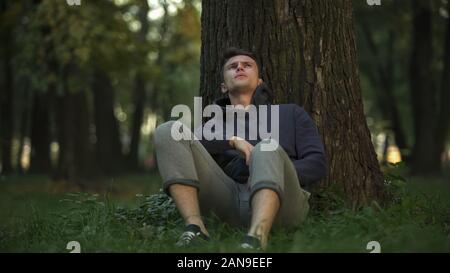 Premuto uomo seduto sotto agli alberi nel parco, problema di disoccupazione, difficoltà Foto Stock