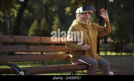 Gioiosa anziana signora utilizzando cuffie vr seduta sul banco in park, intrattenimento Foto Stock