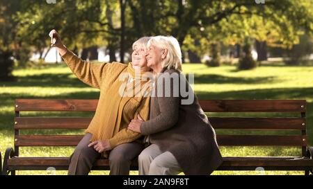 Due vecchie signore prendendo selfie sul telefono cellulare seduta sul banco in park technology Foto Stock