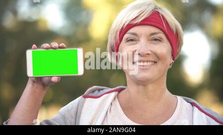 Montare sorridente donna di mezza età che mostra lo schermo verde telefono cellulare, sport app Foto Stock