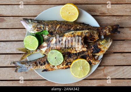 Sani e freschi di pesce di mare, spigola e salpe porgy, piatti alla griglia e servita con succo di limone, lime e foglie di basilico in un colore bianco porcellana piastra sul vecchio tavolo in legno Foto Stock