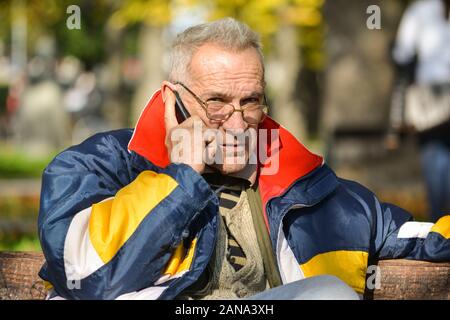 Buona ricerca senior rendendo chiamata telefonica, seduta su una panchina in un parco, godendo di Sun in una luminosa giornata autunnale Foto Stock