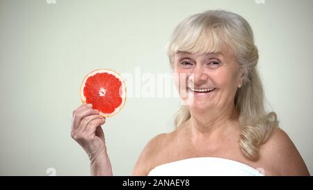 Positivo sorridente donna anziana che mostra fetta di pompelmo alla telecamera, vitamine Foto Stock