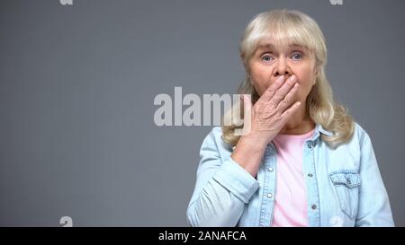 Scioccato invecchiamento della donna che copre la bocca con la mano, stupito con news, emozioni Foto Stock