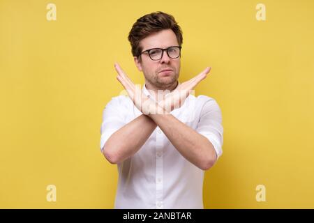 Grave Giovane Uomo in camicia bianca e i bicchieri che mostra il segnale di arresto e guardando la fotocamera. Isolato sfondo giallo Foto Stock