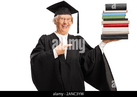 Senior donna in un abito di laurea in possesso di una pila di libri e puntamento isolati su sfondo bianco Foto Stock