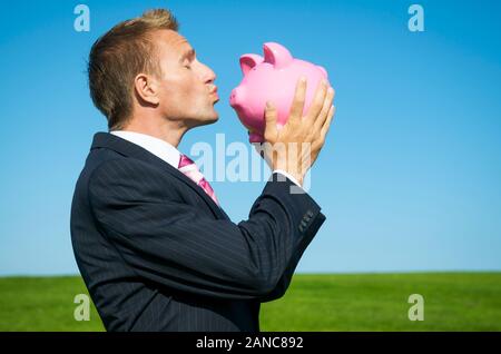 Happy businessman kissing rosa salvadanaio all'aperto sul luminoso verde prato con blue sky spazio di copia Foto Stock