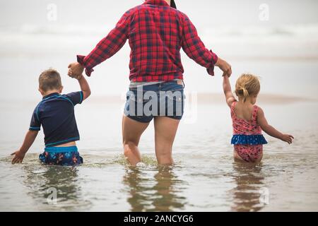 vista posteriore della madre che aiuta i bambini a passare attraverso l'acqua in spiaggia. Foto Stock